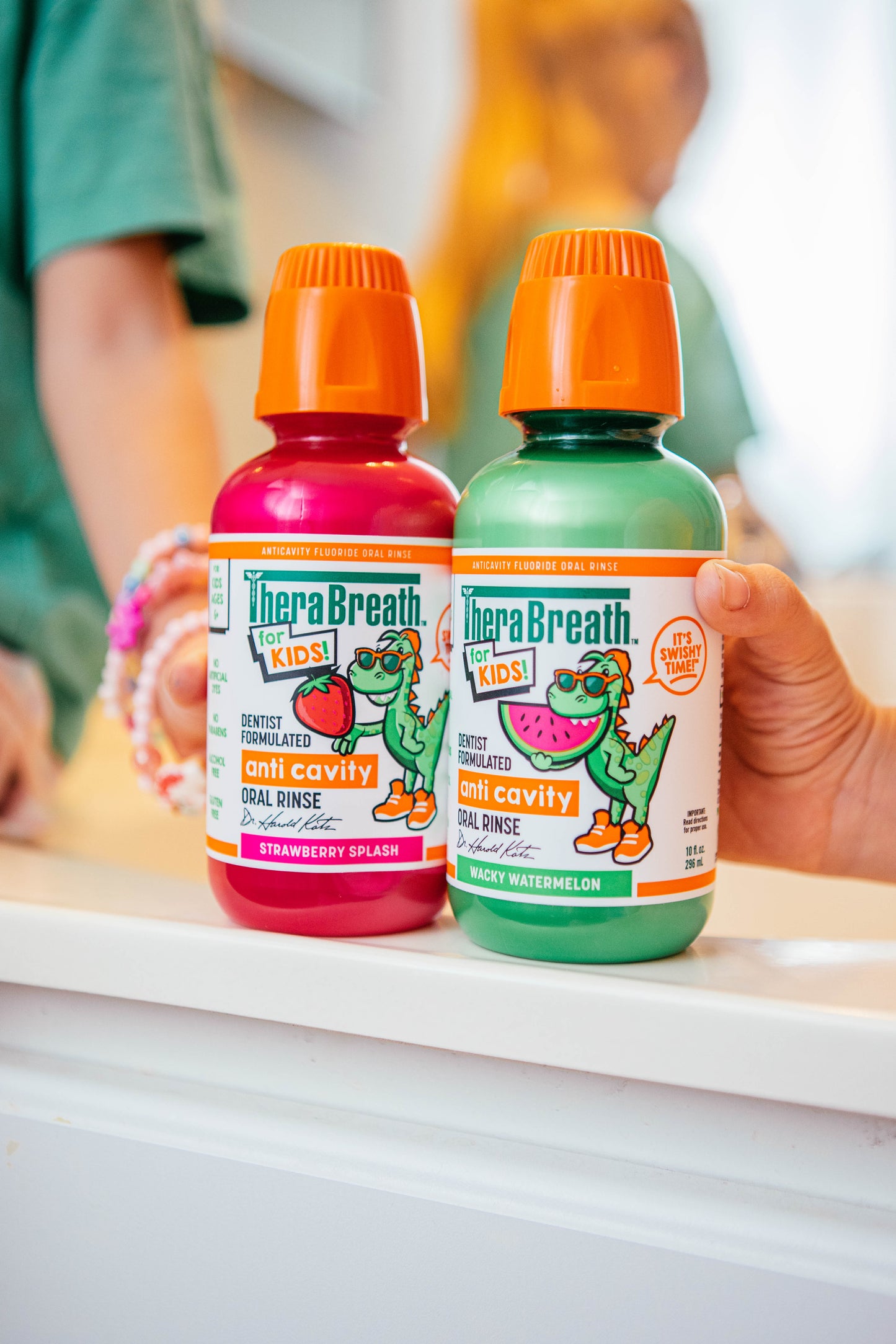 Kids hold two bottles of TheraBreath for Kids Anti-Cavity Oral Rinse: a pink one in Strawberry Splash and a green one in Wacky Watermelon
