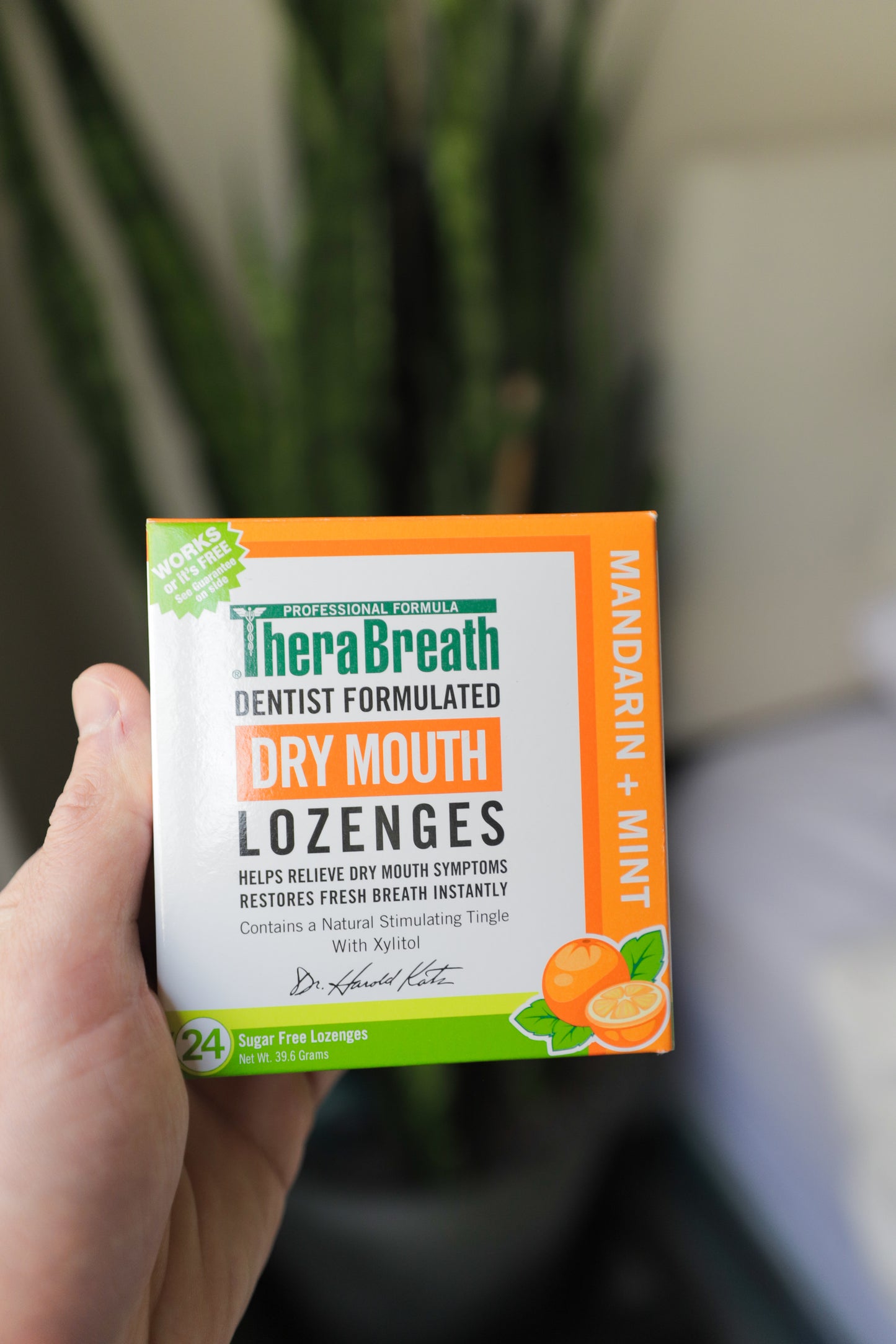 A hand holds a box of TheraBreath Dry Mouth Lozenges in Mandarin Mint. The box is decorated with illustrations oranges and mint leaves.