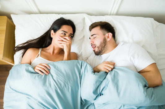 Woman reacting to partner’s bad breath while lying in bed together under a blue blanket