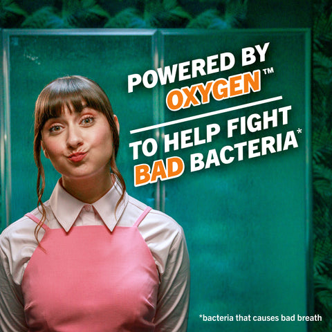 Woman rinsing with TheraBreath Oral Rinse in a green bathroom; powered by oxygen to fight bad‑breath bacteria.